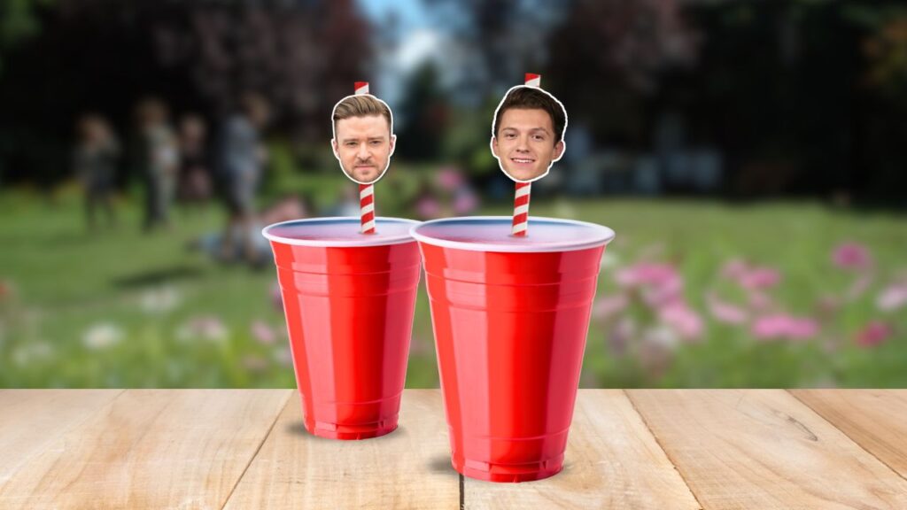 Two red plastic cups with straws featuring celebrity faces of Justin Timberlake and Tom Holland, with a blurred outdoor party scene in the background.