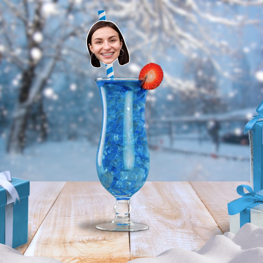 A blue cocktail in a snow-covered winter setting, featuring a personalised face straw with a woman's smiling face and a strawberry garnish.