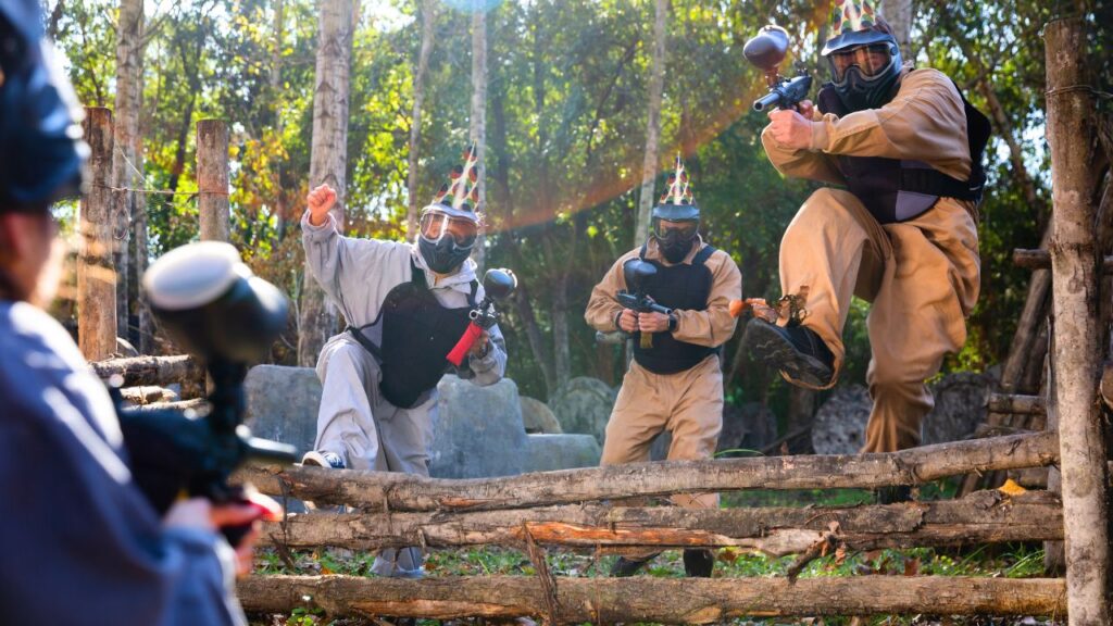 Group of men wearing protective gear and helmets playing paintball in a forest setting as part of a stag do activity.